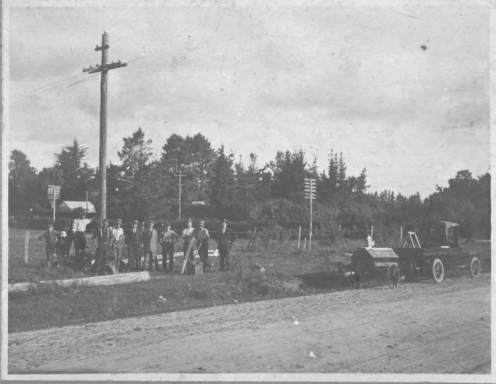 Group photo of 10 Power Board men and two children on side of road with Cambridge Power Board vehicle and trailer on right. Early 1920s
